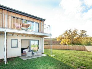 A modern two-story house with a balcony, a patio with chairs, and a large tree in the background.
