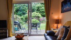 A living room with a sofa, a coffee table with a fruit bowl, a TV, and a sliding glass door leading to a garden with outdoor furniture. - Between the forest and the sea (New Milton, near Lymington)