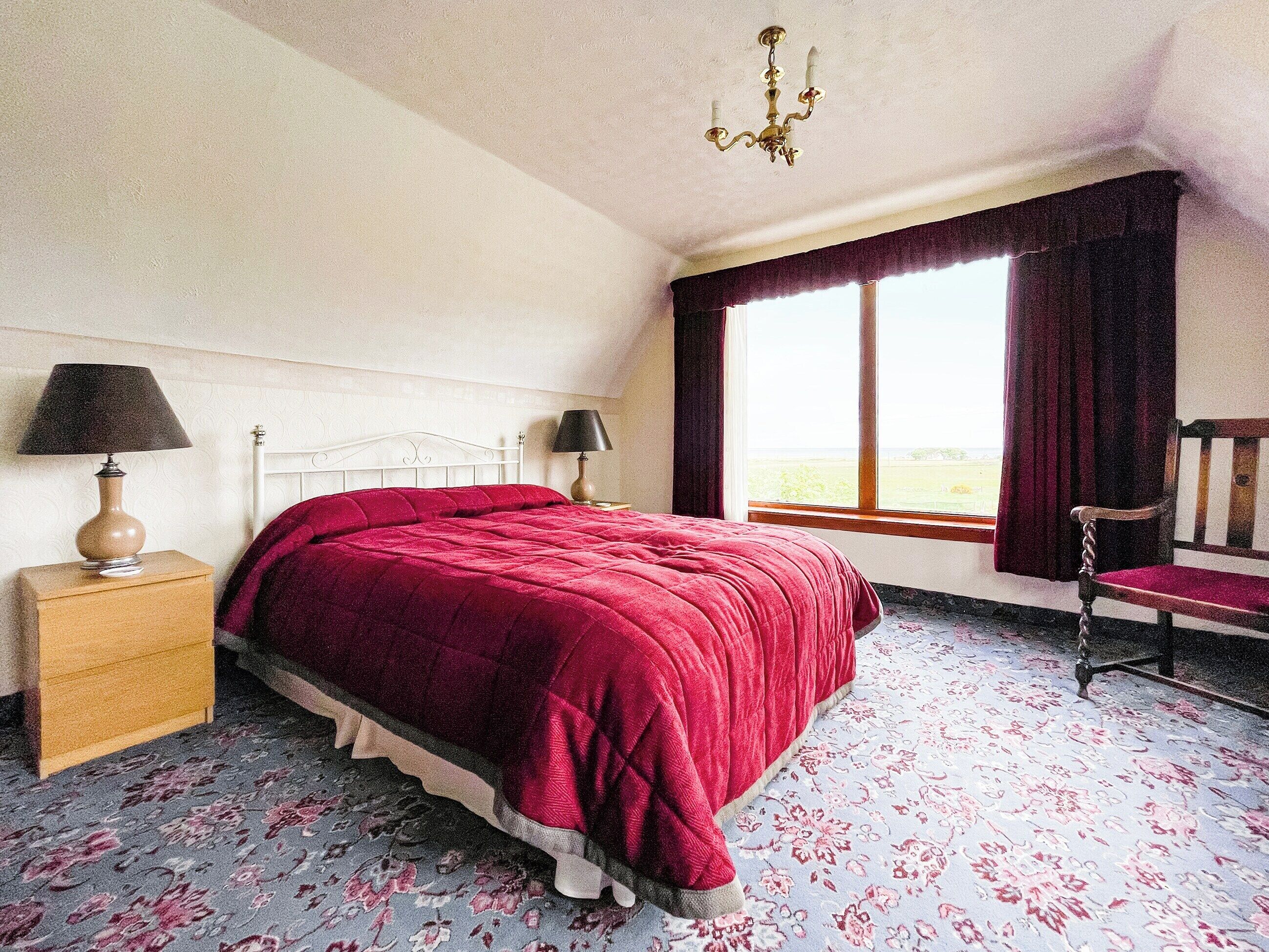 Un dormitorio con una cama grande, una mesita de noche con lámpara, una silla y una ventana con cortinas.
