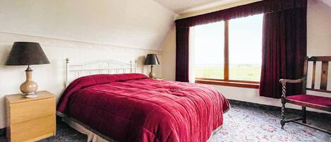 Un dormitorio con una cama grande, una mesita de noche con lámpara, una silla y una ventana con cortinas.