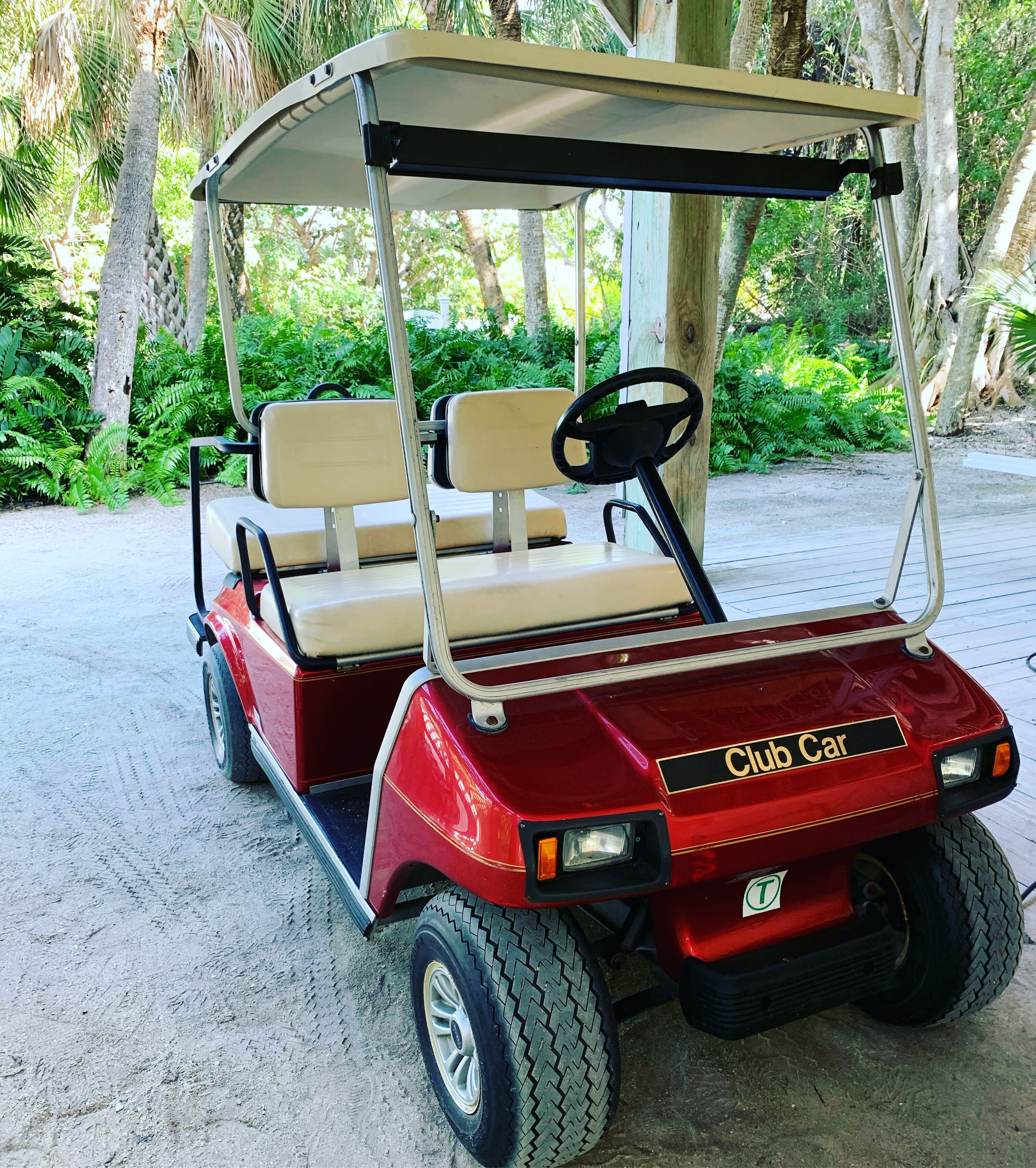 Carro de golf rojo con toldo, identificado como 'Club Car', estacionado en un camino arenoso rodeado de follaje.