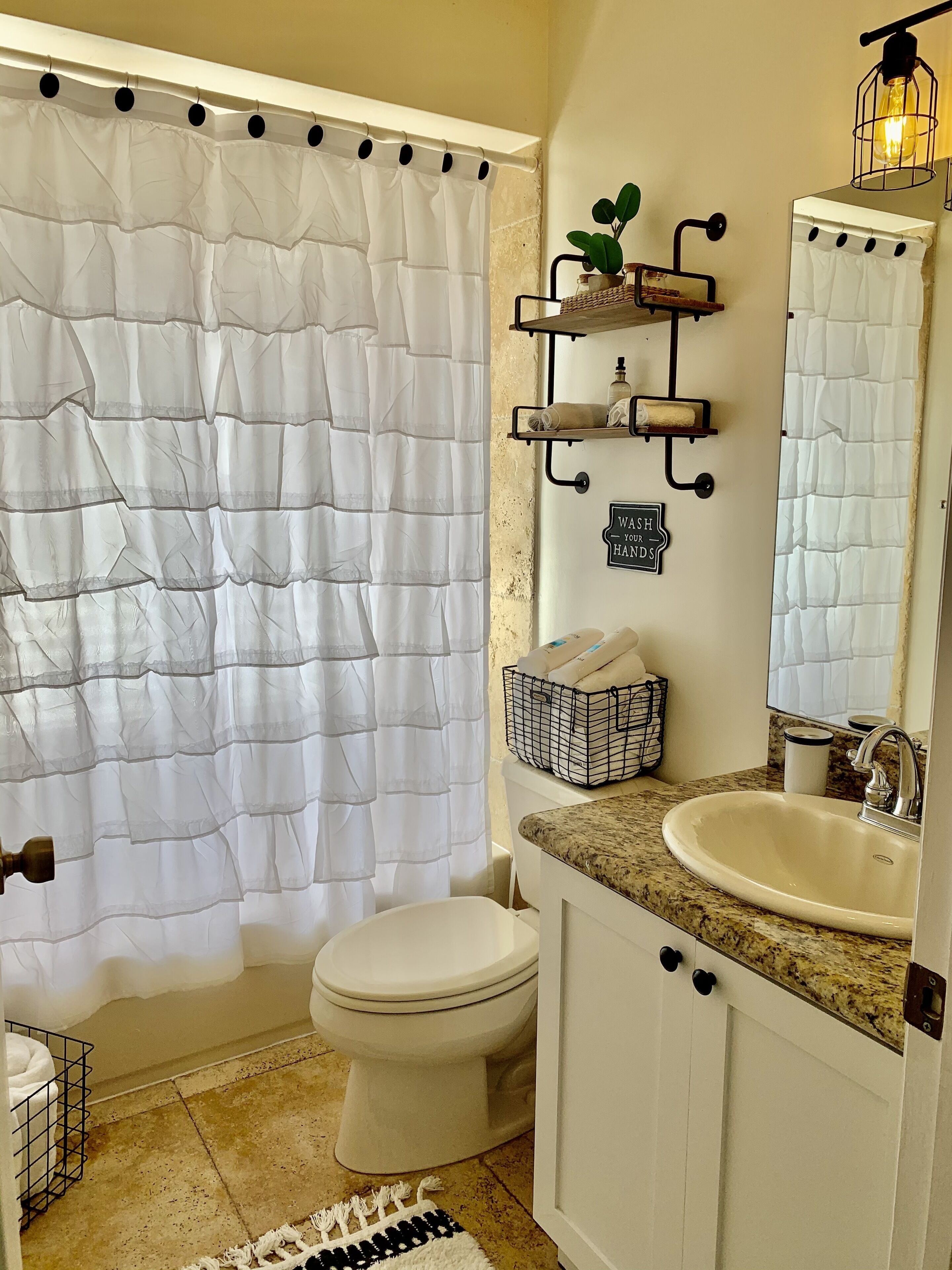 Un baño con cortina de ducha blanca, inodoro, lavabo con encimera de granito y repisa con artículos de aseo.