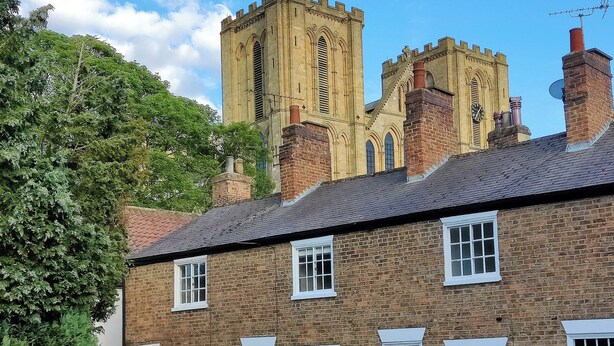 A traditional brick building with multiple windows and doors, a chimney, and a large tower in the background. - 2 bedroom accommodation in Ripon (Ripon)