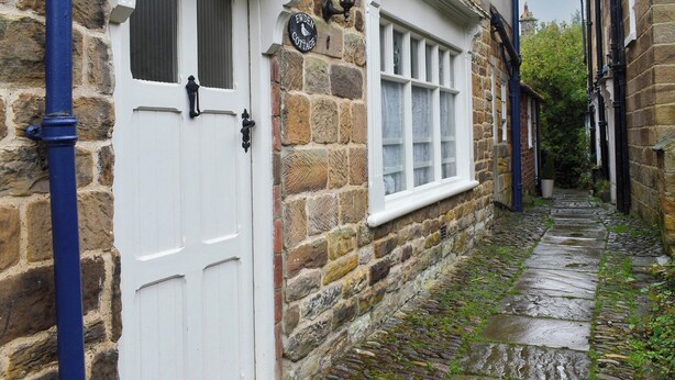 Uma estreita viela de pedra com uma porta e janela brancas, uma placa com a inscrição "EWDEN COTTAGE" e um cano azul.