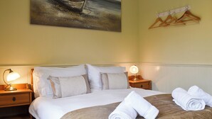 Cama bem arrumada com lençóis brancos, duas luminárias de cabeceira, um quadro de um barco na parede e um cabideiro com ganchos.