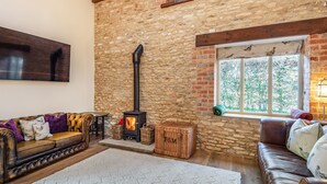 A cozy living room with a stone wall, a fireplace, leather sofas, a flat-screen TV, and a large window with a view of trees.