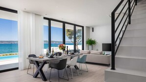 Una sala de estar moderna con comedor, ventanas amplias y vista al mar, una escalera y un televisor de pantalla plana.