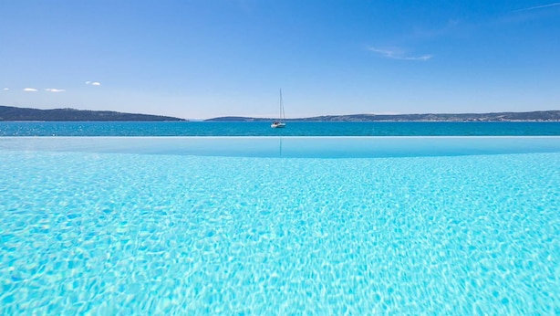 Una piscina azul cristalina que se extiende hasta un mar tranquilo con un velero a lo lejos.