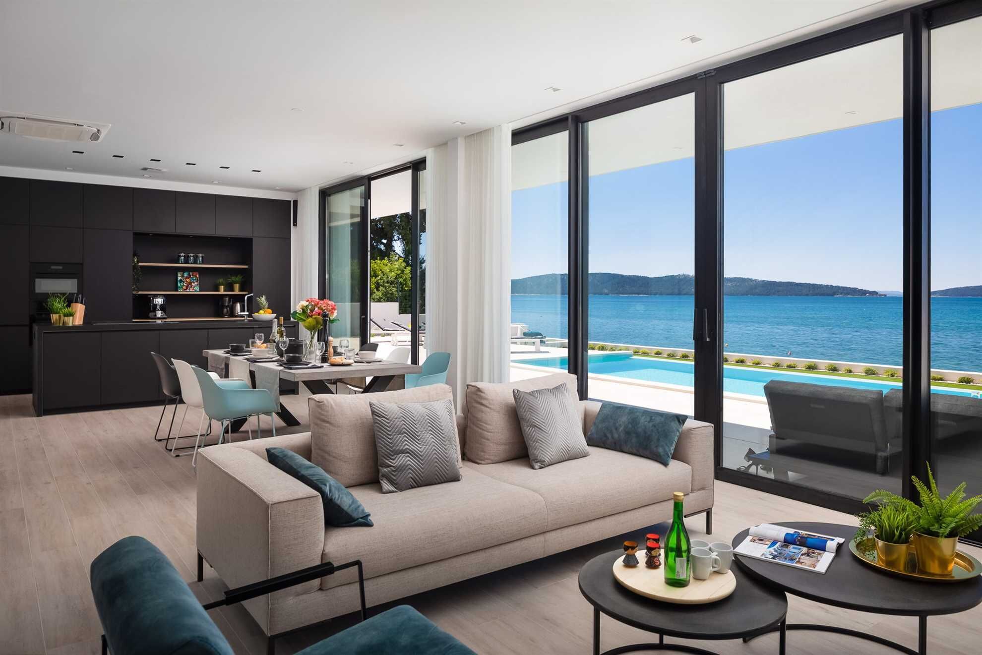 A modern living room with a sofa, coffee table, and dining area. There is a large window with a view of the sea.