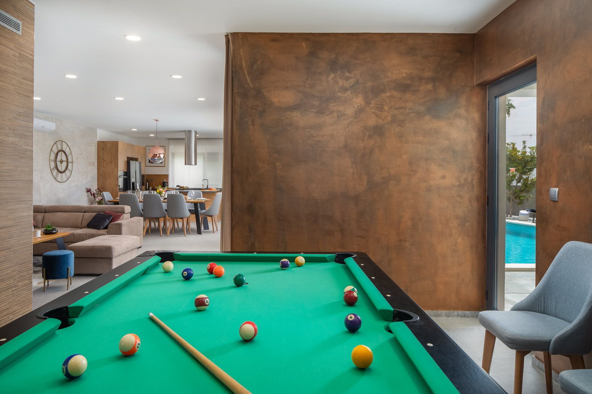 Una mesa de billar en una habitación moderna con zona de comedor y vista a una piscina.