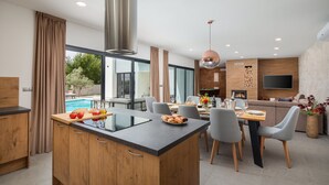 Een moderne keuken met een centraal eiland, een eetgedeelte met grijze stoelen, een groot raam en een zwembad buiten.