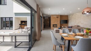 Een moderne woonkamer met een open haard, een eethoek met een tafel gedekt voor twee personen, en een groot raam met uitzicht op een zwembad.