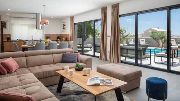 Een moderne woonkamer met een ruime hoekbank, een houten salontafel en uitzicht op een zwembad en stadsbeeld door grote ramen.