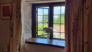Une pièce avec une fenêtre donnant sur un mur blanc et une étagère en bois. Un petit vase avec des fleurs est posé sur l’étagère.