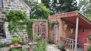 Un bâtiment en pierre avec une porte en bois, une construction en briques avec une porte rouge, et un petit jardin agrémenté de diverses plantes et fleurs.