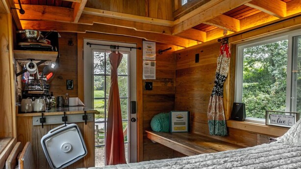 A cozy cabin interior with a bed, a small kitchenette, a window with a view of greenery, and a curtain.