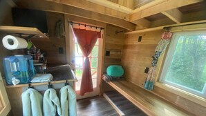 A compact, wooden interior with a small kitchenette, a window with a view of trees, and a door with a red curtain.