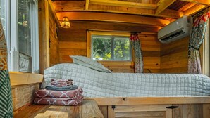 A cozy cabin room with a bed, a window with curtains, a wall-mounted air conditioner, and a small table with towels.