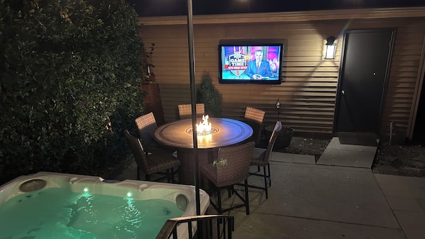 An outdoor patio with a hot tub, a round table with chairs, and a television screen displaying a news program. - PRIME Location with Hot Tub, Pool Table and Sauna (Columbus)