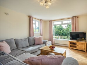 A living room with a grey sofa, a wooden coffee table with fruit, a TV on a wooden stand, and large windows with curtains. - 3 bedroom accommodation in Hundred House, near Llandrindod Wells (Hundred House, near Llandrindod Wells)