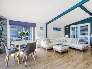 A modern living room with a white sofa, a dining table with chairs, and a large window with blinds. - Norfolk Broads and Coast, The Beach House (Winterton on Sea)