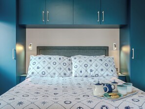 Un lit avec une couvre-lit aux motifs marins, une table de chevet avec une théière et des tasses, et des armoires de couleur turquoise avec des poignées.