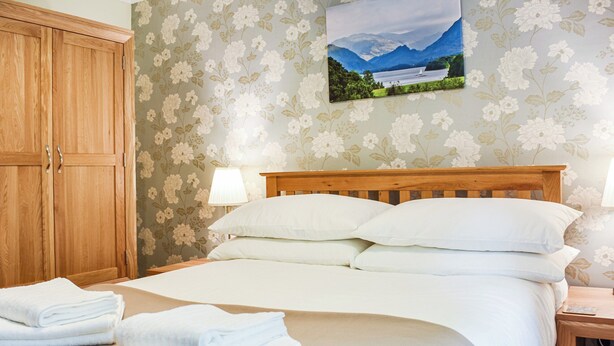 A neatly made bed with white linens and pillows, a wooden headboard, a wooden wardrobe, and a floral wallpaper. - 3 bedroom accommodation in Keswick (Keswick)