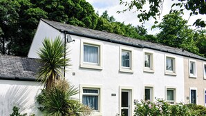 A white two-story building with multiple windows and a door, surrounded by greenery and a small garden. - 3 bedroom accommodation in Keswick (Keswick)