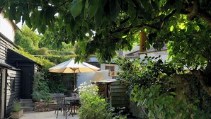 A patio area with a table and chairs under a large umbrella, surrounded by lush greenery and a stone wall. - Church View Cottage & Barn (Hatherleigh, near Okehampton)