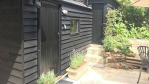 A black wooden building with a door, a small window, and a porch area with steps and potted plants. - Church View Cottage & Barn (Hatherleigh, near Okehampton)