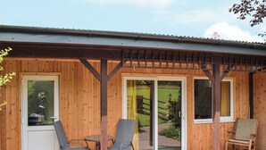 Een houten gebouw met een overdekt terras, twee tafels met stoelen, een barbecue en uitzicht op een gazon.