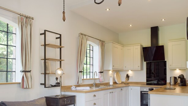 A modern kitchen with white cabinets, a wooden countertop, and a built-in oven. There is a window with curtains, a small table with a lamp, and a sofa with cushions.