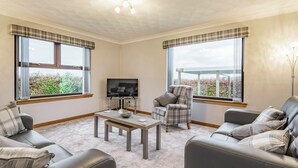 Una sala de estar con dos sofás, una mesa de centro, un televisor y una silla. Amplias ventanas con cortinas y vistas al exterior, con zonas verdes.