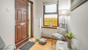 Un recibidor acogedor con una puerta de madera, un ventanal con vistas al follaje otoñal, una silla con cojín y una mesita con un jarrón de flores.