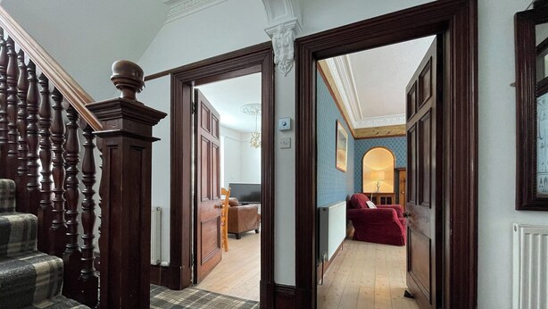 Un couloir avec un escalier en bois, une moquette à carreaux, un canapé rouge et un téléviseur à l’arrière-plan.