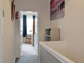 Un pasillo con piso alfombrado, un sillón mecedor de madera, una estantería y una puerta que da a otra habitación.