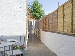 A narrow outdoor patio with a round table and chairs, a white brick wall, and a wooden fence.
