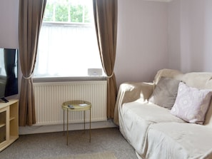 A room with a beige sofa, a small round table, a TV on a stand, and a window with curtains.