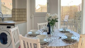 A dining area with a table set for four, surrounded by white chairs, a washing machine, and a window with blinds. - 3 bedroom accommodation in Buxton (Buxton)