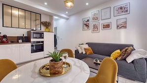 Une cuisine moderne avec des meubles blancs, un four encastré et une machine à café. On y trouve un coin repas avec une table ronde, des chaises et une plante en pot. Un canapé gris avec des coussins se trouve dans le salon, et des tableaux encadrés ornent le mur.