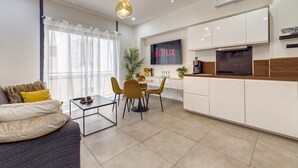 Un salon moderne comprenant un canapé, une table basse et un coin repas avec des chaises. On y trouve également une cuisine avec des meubles blancs, un téléviseur et une machine à café.