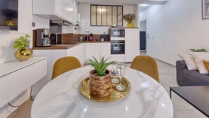 Une cuisine moderne avec des meubles blancs, un four encastré et une machine à café. On y trouve également un coin repas avec une table et des chaises en marbre.