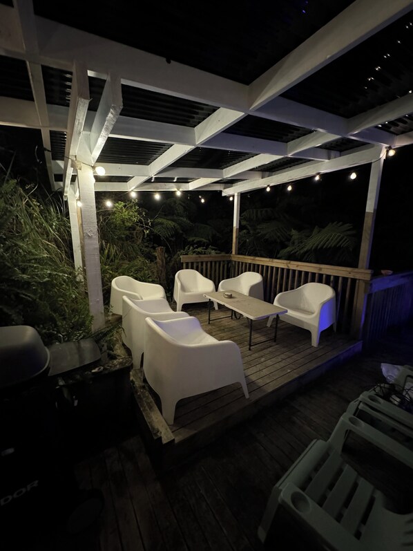 An outdoor patio with white plastic chairs and a table, surrounded by wooden decking and greenery.