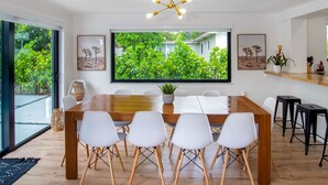 A modern dining area with a large wooden table, white chairs, and a large window offering a view of greenery outside. - Boho Chic House-POOL & FIREPIT - 15 mins to South Beach (El Portal)