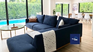 A modern living room with a dark blue sofa, a glass coffee table, and a view of a swimming pool and greenery outside. - Boho Chic House-POOL & FIREPIT - 15 mins to South Beach (El Portal)