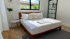 A bedroom with a wooden bed, white bedding, a small bedside table, a hanging plant, and a view of the outside through a window. - Boho Chic House-POOL & FIREPIT - 15 mins to South Beach (El Portal)