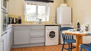 A compact kitchen with a washing machine, a refrigerator, and a dining area with a table and chairs. - The Cottage - UK35177 (Buckland Brewer, near Bideford)