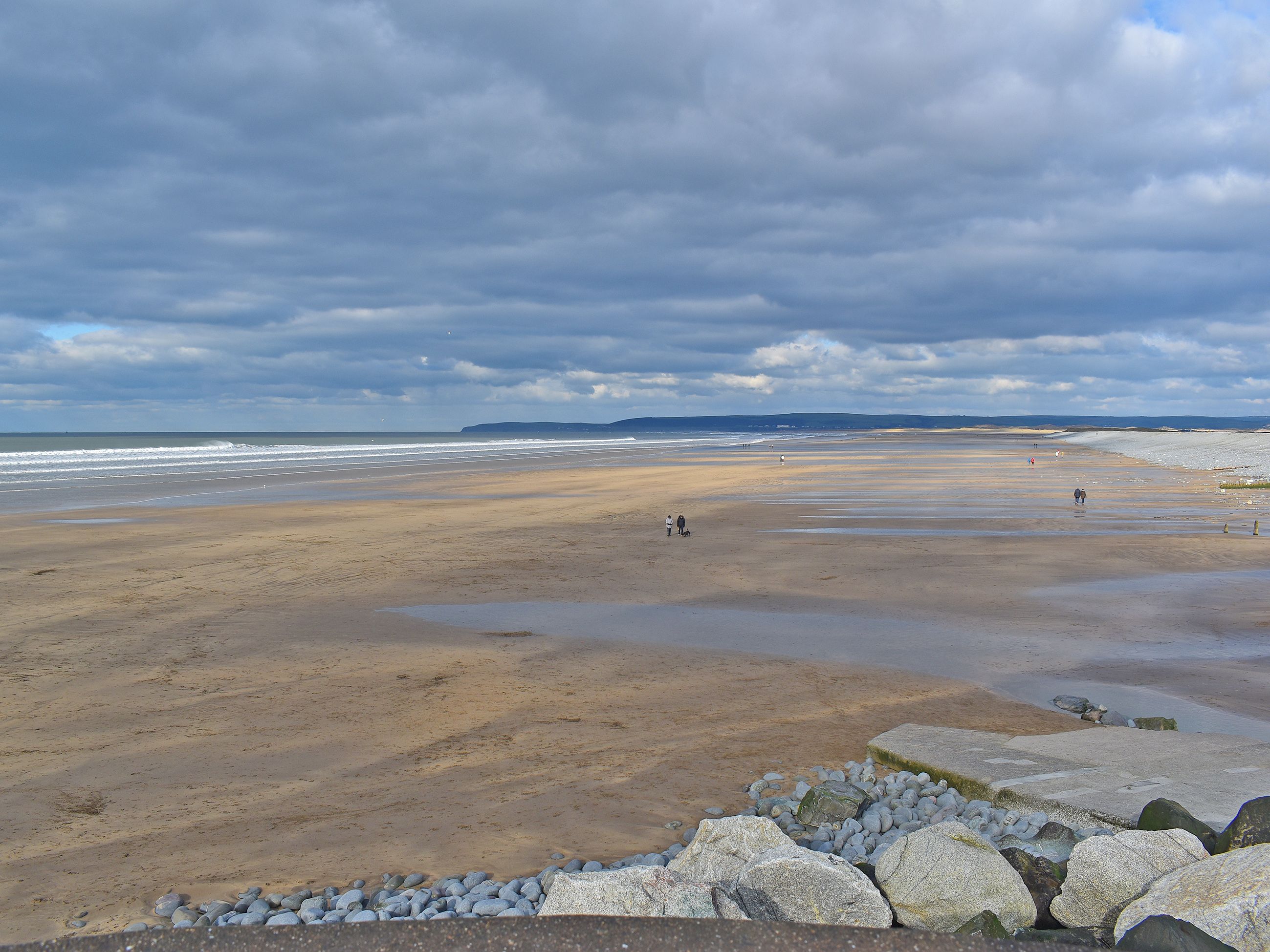 Ein weiter Sandstrand mit steiniger Uferlinie und einem fernen Blick auf das Meer unter bewölktem Himmel.