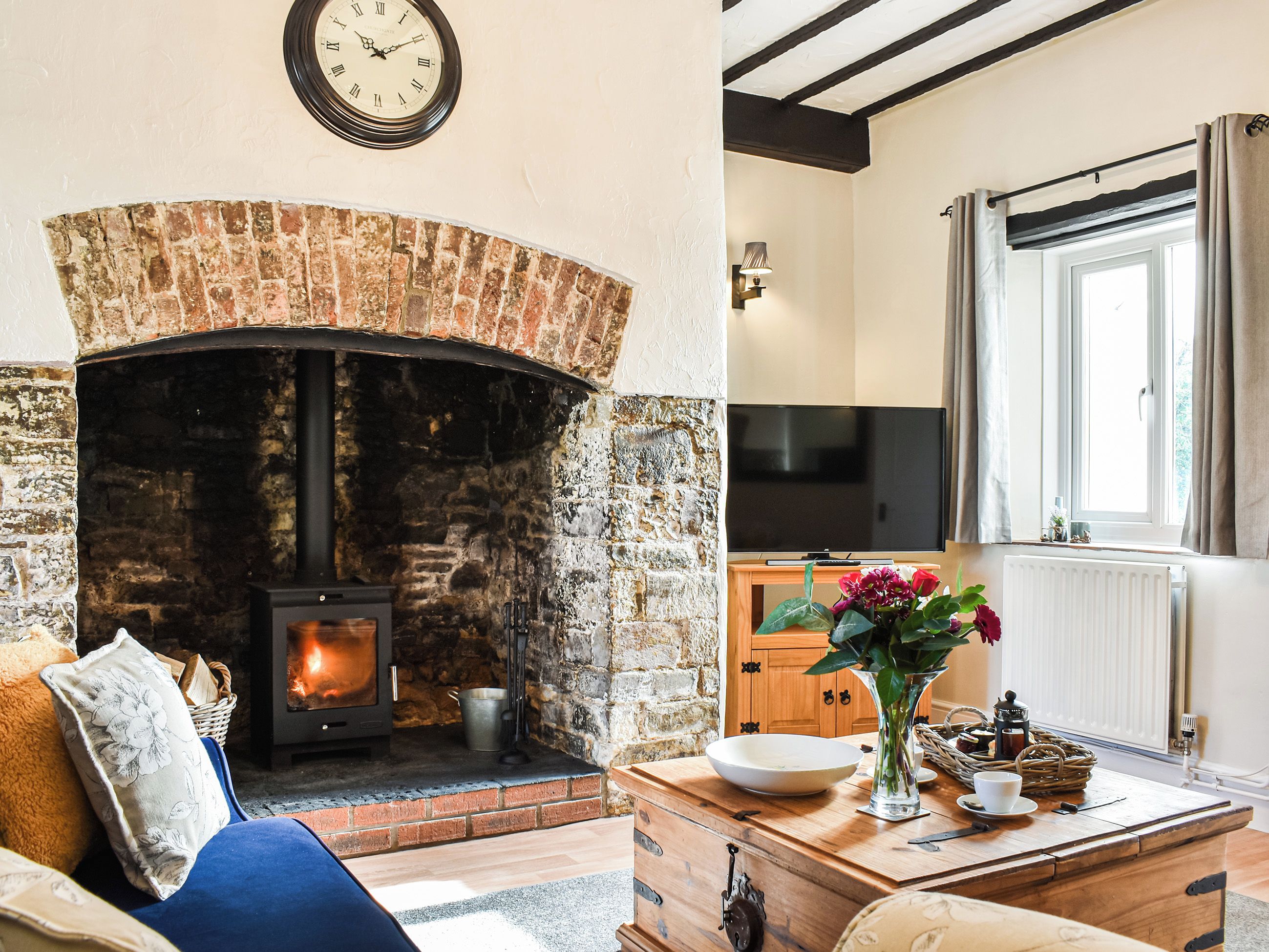 A cozy living room with a brick fireplace, a television, and a dining area with a vase of flowers.