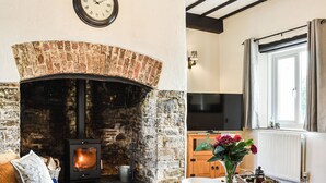 A cozy living room with a brick fireplace, a television, and a dining area with a vase of flowers. - The Cottage - UK35177 (Buckland Brewer, near Bideford)